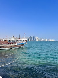 “Traditional wooden dhow boat moored along the corniche in Doha, Qatar, with the modern skyline of West Bay in the background. Clear blue skies and turquoise waters of the Arabian Gulf create a vibrant contrast, highlighting the blend of cultural heritage and contemporary architecture