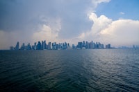 A panoramic view of Doha’s modern skyline from across the Arabian Gulf, with towering skyscrapers set against a cloudy sky.