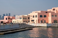 This image shows the colorful pastel buildings of Old Doha Port, a vibrant waterfront district in Qatar. The buildings feature modern designs with artistic murals, set against the backdrop of Doha’s skyline. The waterfront promenade and steps lead down to the water, creating a lively yet serene atmosphere.